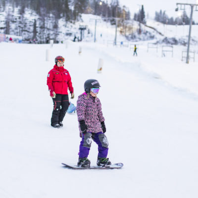 Lapsi opettelee lumilautailua rinteessä hiihdonopettajan kanssa.